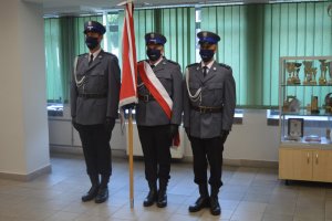 Trzech funkcjonariuszy stojących obok siebie w szeregu, środkowy funkcjonariusz trzyma sztandar Komendy Powiatowej Policji w Nowym Targu
