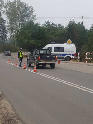 Policjant ruchu drogowego (widoczna biała czapka, żółta kamizelka) kontroluje samochód zatrzymany na drodze, na wjeździe do strefy nadgranicznej. Pora dzienna.