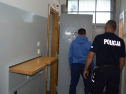 zatrzymany w niebieskiej bluzie stoi przed otwartymi drzwiami pomieszczenia dla osób zatrzymanych a obok stoi policjant