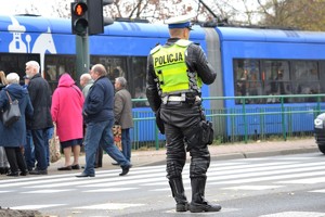 Policjant ruchu drogowego ubrany w skórzany strój motocyklowy kieruje ruchem na przejściu dla pieszych. Stoi przodem do pojazdów przepuszczając pieszych.