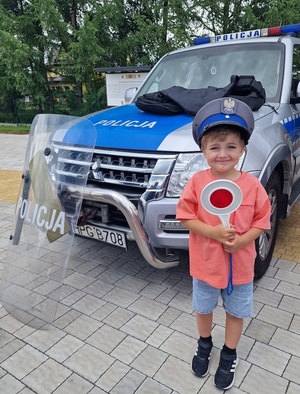 dziecko stoi przed radiowozem w policyjnej czapce i ma lizak w dłoni
