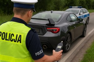 Policjant z sonometrem w ręce oraz w tle radiowóz z kontrolowanymn pojazdem